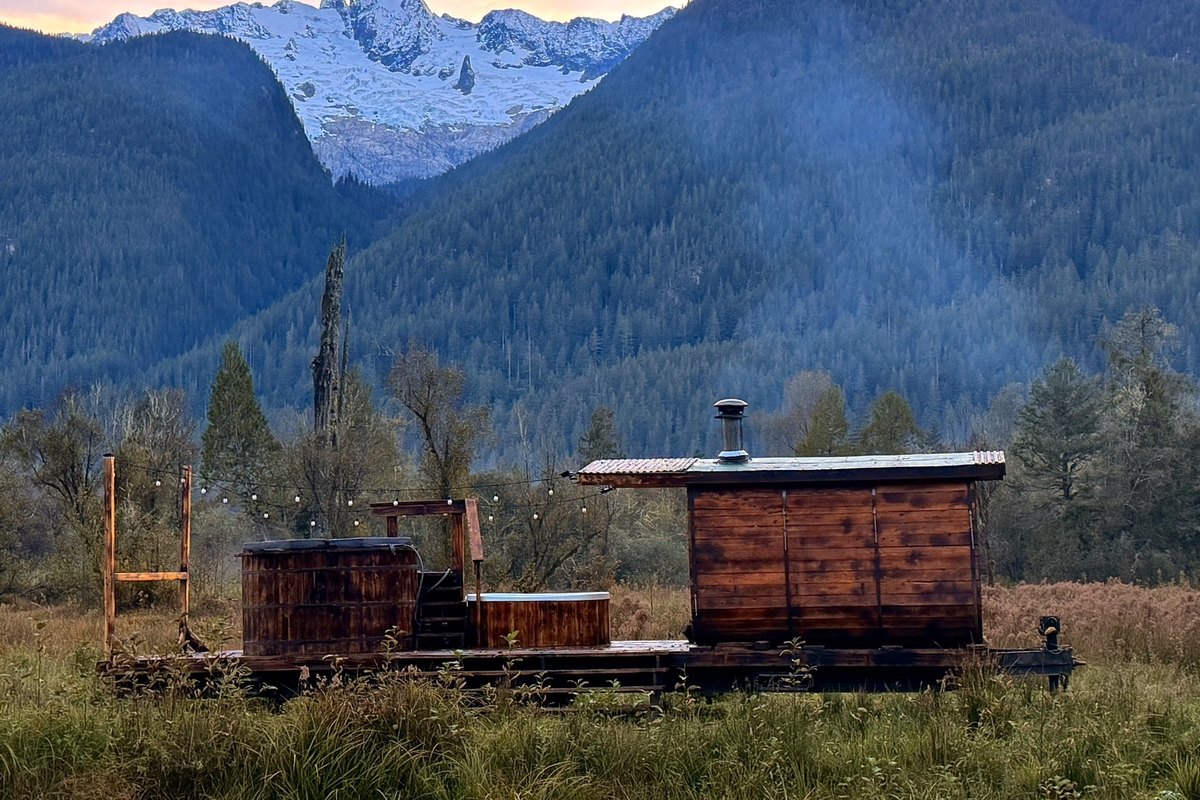 Alpha sauna with wood-fired hot tub and cold plunge in a meadow below snowy mountains at sunset