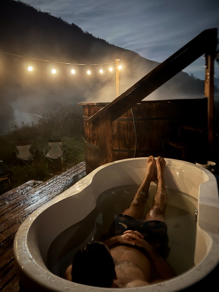 Guest relaxing in the cold plunge beside the steaming wood-fired hot tub under string lights